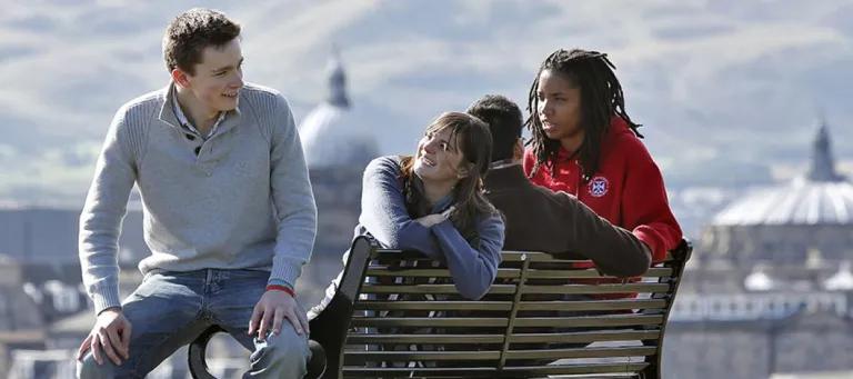 University of Edinburgh students sit on a bench with Edinburgh skyline behind them