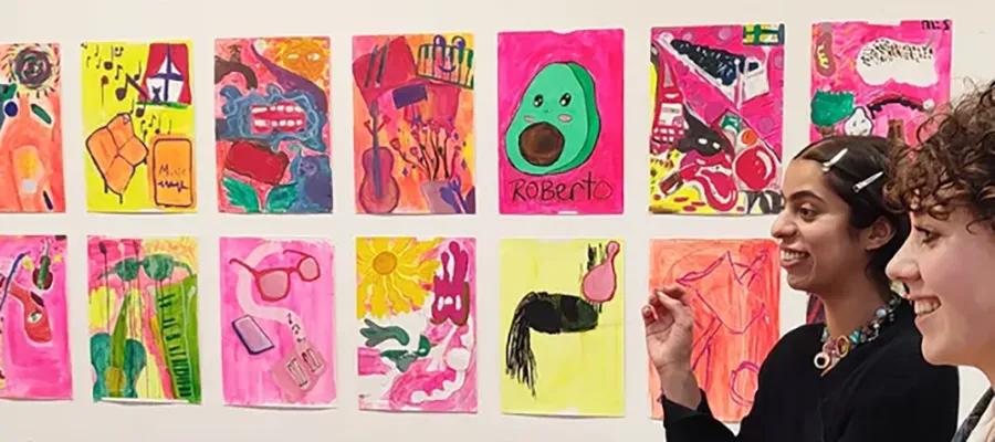 Two people standing and smiling in front of a wall filled with 14 colourful artworks. 