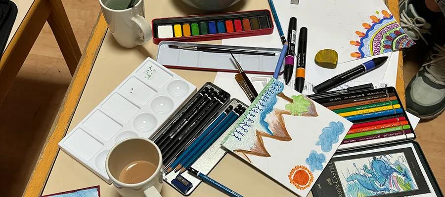 A table covered in sketchbooks, pencils, paint and mugs.